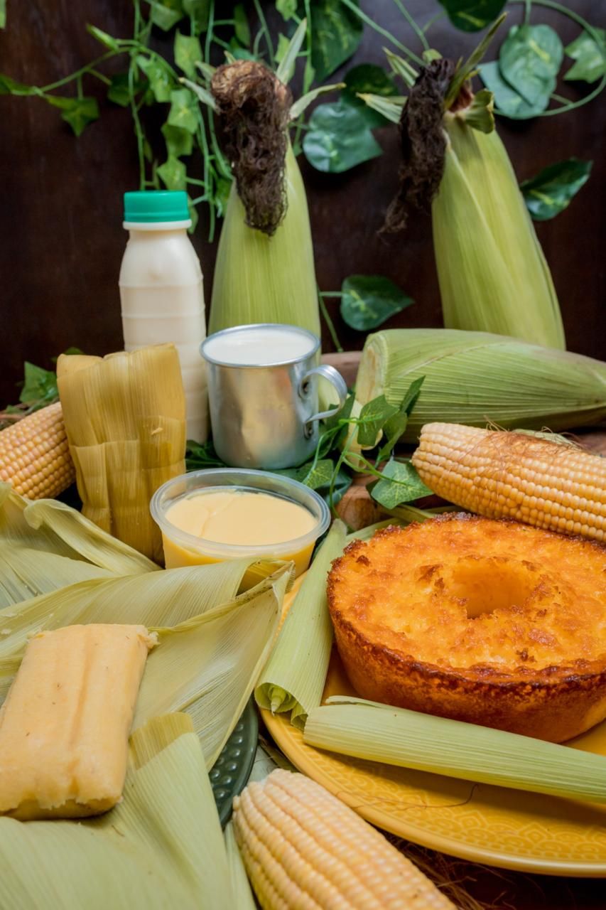 Pamonhas, bolo de milho, curau e garrafa de leite entre espigas de milho verde